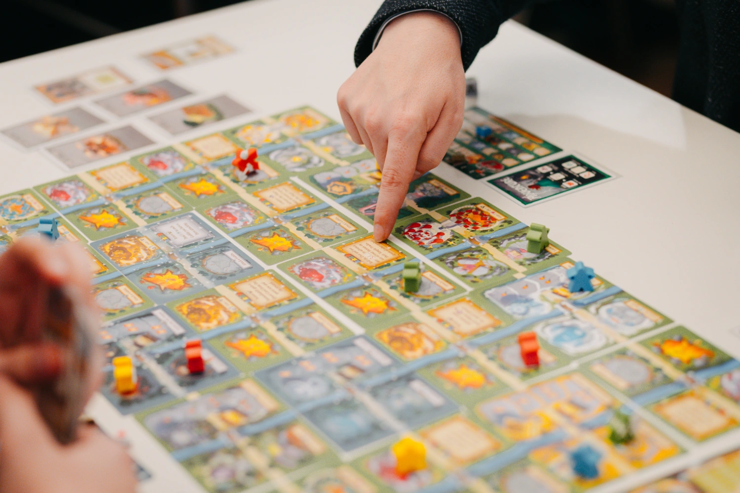Person zeigt mit Zeigefinger auf ein Spielfeld am Tisch mit vielen bunten Kärtchen in einem Raster angeordnet und Figuren darauf.