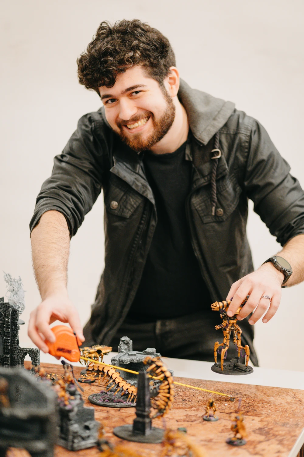 Mann lächelt freundlich in die Kamera und hält dabei ein Maßband und eine Warhammer-Miniaturenfigur in der Hand, die er gleich auf dem Spielfeld vor sich platzieren wird.