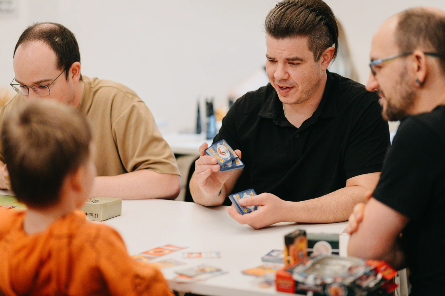 Erwachsene Personen und ein Kind sitzen um einen Tisch und spielen Pokémon TCG.