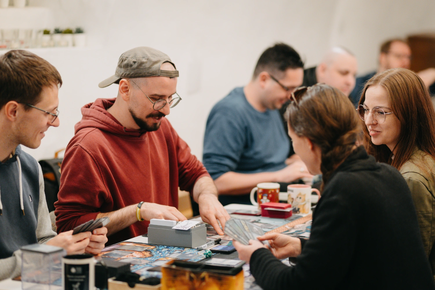 Frauen und Männer sitzen lachend um einen Tisch und spielen ein Sammelkartenspiel.