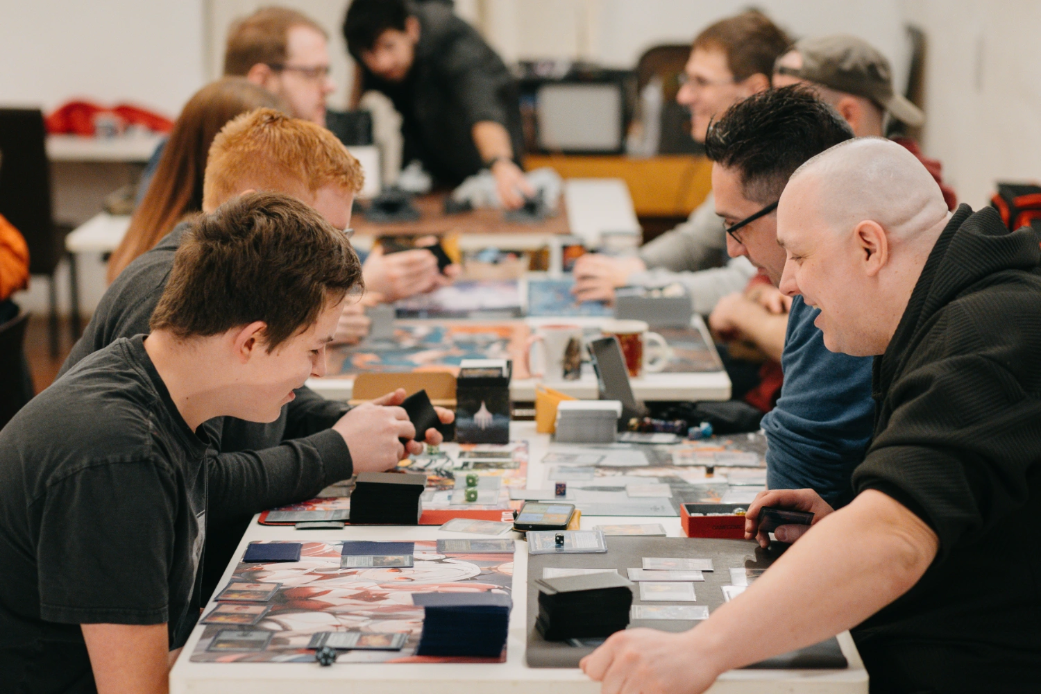 Vier Personen um einen Tisch, die auf das vor sich liegende Spielfeld schauen und ein Sammelkartenspiel spielen.