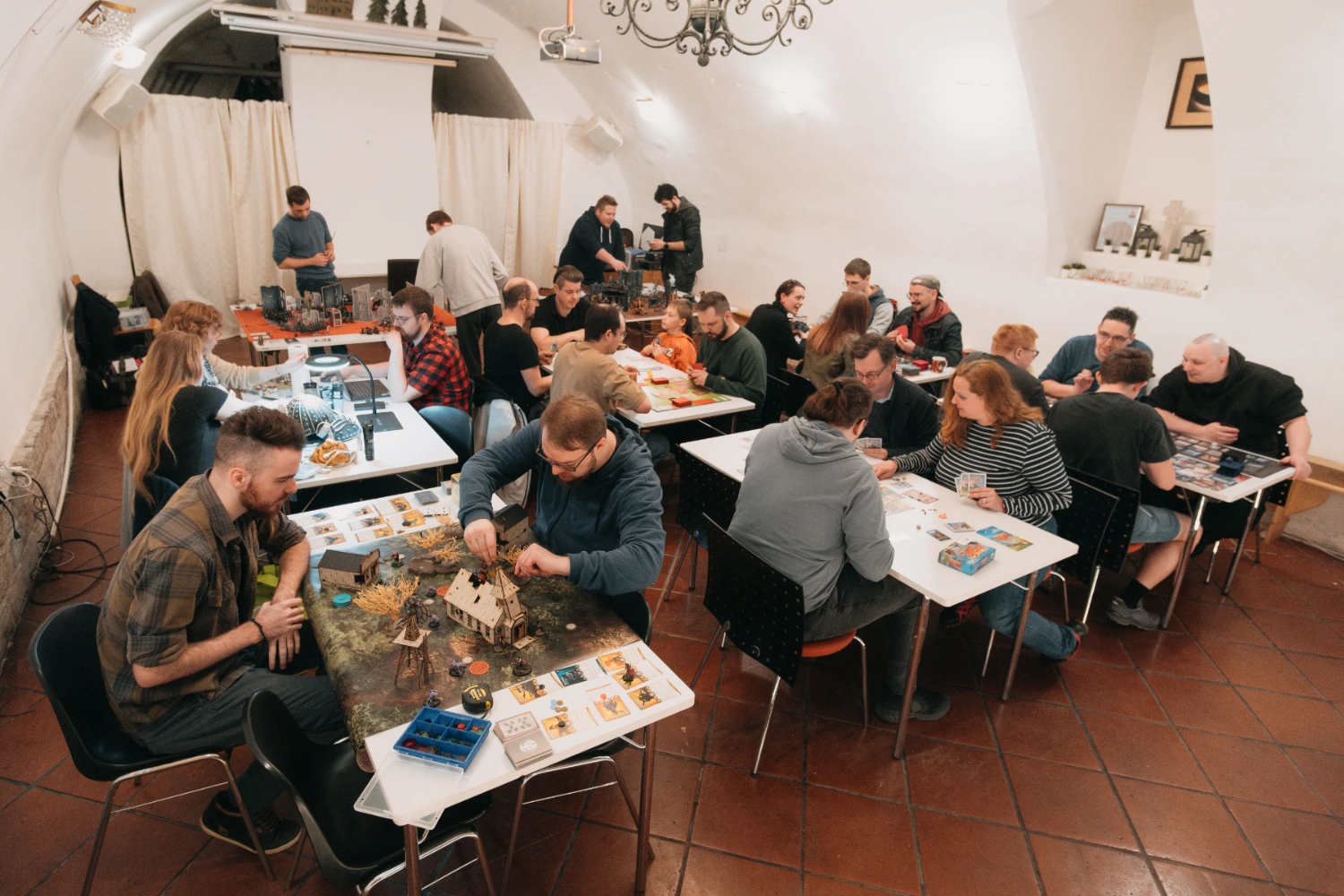 Viele Personen sitzen in einem Raum um Tische herum und spielen unterschiedliche Arten von Spielen.